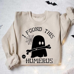 May include: A beige sweatshirt with a black graphic of a ghost holding a bone with the text "I found this humerus".