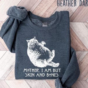 May include: Heather dark gray sweatshirt with a white graphic of a cat lying on its back. The text "MOTHER, I AM BUT SKIN AND BONES" is below the cat. The sweatshirt is folded on a wooden surface.