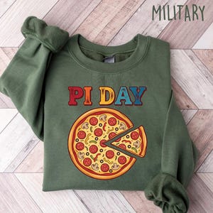 May include: Military green sweatshirt with the text "PI DAY" in red and blue letters above a pizza graphic. The pizza has a slice cut out, showing toppings like pepperoni and mushrooms. The sweatshirt is folded on a light wood-grain surface.