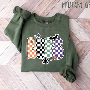 May include: A green sweatshirt with the word "Boo" written in orange, black, and green checkered letters. The letters are outlined in black and have a white background. There are also small black bats, stars, and a spider on the sweatshirt.