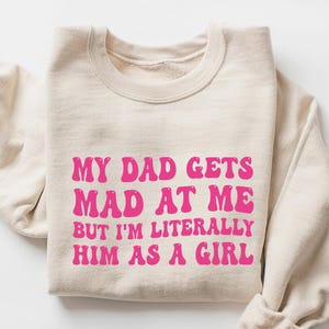May include: A sand-colored sweatshirt with the text "MY DAD GETS MAD AT ME BUT I'M LITERALLY HIM AS A GIRL" in bright pink, retro-style lettering. The sleeves are folded, and the garment is displayed on a white surface.