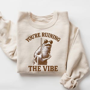 May include: Sand-colored sweatshirt with the text "YOU'RE RUINING THE VIBE" and a frog wearing sunglasses. The frog is holding a pole. The word "SAND" is in the upper right corner.