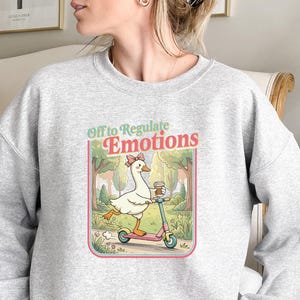 May include: A light gray sweatshirt featuring a cartoon goose riding a pink scooter and holding a coffee cup. The graphic includes the text "Off to Regulate Emotions" in a retro style. The goose wears a bow and the background is a park scene.