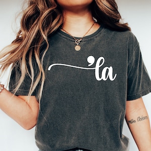 May include: A dark gray t-shirt with the word "'la" written in white cursive lettering. The shirt is being worn by a person with long brown hair and a gold necklace.