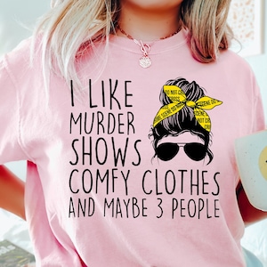 May include: Pink t-shirt with a black graphic that says "I like murder shows, comfy clothes and maybe 3 people". The graphic also includes a woman's hair in a bun with sunglasses and yellow crime scene tape.