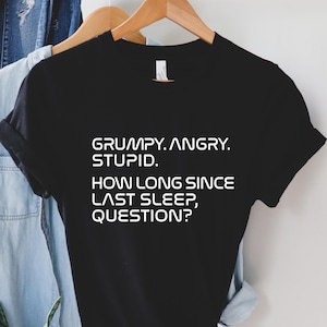 May include: Black t-shirt with white text: "GRUMPY. ANGRY. STUPID. HOW LONG SINCE LAST SLEEP, QUESTION?" The shirt is hanging on a wooden hanger with a light blue denim shirt and a brown hat in the background.