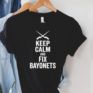 May include: Black t-shirt with the white text "KEEP CALM AND FIX BAYONETS" and crossed rifles. The shirt is hanging on a wooden hanger with a light blue denim shirt and a brown hat in the background.