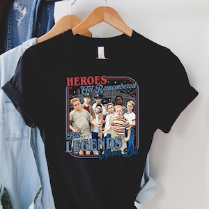 May include: Black t-shirt featuring a graphic of a group of children with the text "HEROES Get Remembered But LEGENDS Never Die" in red and blue. The shirt is hanging on a wooden hanger with other clothing.