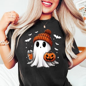 May include: A black t-shirt with a white ghost wearing an orange and brown knit hat holding a pumpkin and a cup of iced coffee. The ghost has a cute face with big eyes and a small mouth. There are bats and stars in the background.