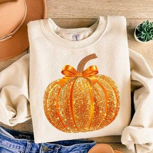 May include: A cream colored sweatshirt with a sparkly gold pumpkin with a brown stem and a gold bow. The pumpkin is centered on the sweatshirt.