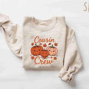 May include: A sand-colored crewneck sweatshirt featuring a graphic of three smiling pumpkins in orange and light pink, surrounded by small autumn leaves. The text "Cousin Crew" is printed in orange above and below the pumpkin illustration.