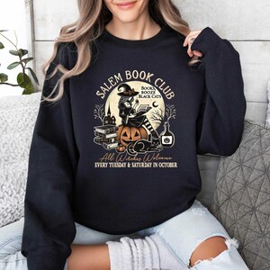May include: Black sweatshirt with a graphic of a witch reading a book in front of a pumpkin. The text reads "Salem Book Club" with the words "Books, Booze, Black Cats" in a smaller font. The text at the bottom reads "All Witches Welcome Every Tuesday & Saturday in October."