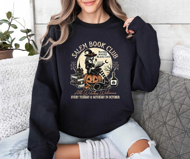 May include: Black sweatshirt with a graphic of a witch reading a book in front of a pumpkin. The text reads "Salem Book Club" with the words "Books, Booze, Black Cats" in a smaller font. The text at the bottom reads "All Witches Welcome Every Tuesday & Saturday in October."