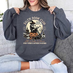 May include: A dark gray sweatshirt with a graphic design of a witch reading a book in front of a pumpkin. The text on the graphic says "Salem Book Club" and "Books Booze Black Cats". The text at the bottom says "All Witches Welcome Every Tuesday & Saturday in October".
