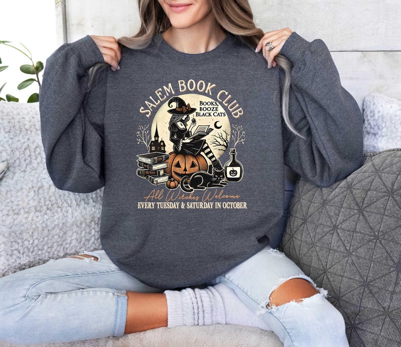 May include: A dark gray sweatshirt with a graphic design of a witch reading a book in front of a pumpkin. The text on the graphic says "Salem Book Club" and "Books Booze Black Cats". The text at the bottom says "All Witches Welcome Every Tuesday & Saturday in October".