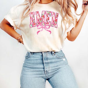 May include: A white t-shirt with a floral design that spells out the word "AMEN" in pink letters with a pink bow underneath.
