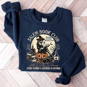 May include: Navy blue sweatshirt with a graphic of a witch reading a book in front of a pumpkin and the text "Salem Book Club" with the words "Books, Booze, Black Cats" underneath. The text at the bottom reads "All Witches Welcome Every Tuesday & Saturday in October."