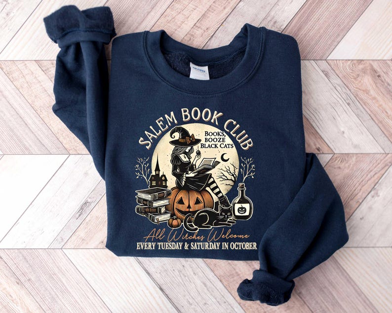 May include: Navy blue sweatshirt with a graphic of a witch reading a book in front of a pumpkin and the text "Salem Book Club" with the words "Books, Booze, Black Cats" underneath. The text at the bottom reads "All Witches Welcome Every Tuesday & Saturday in October."