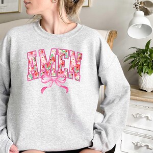 May include: A gray sweatshirt with a floral print of the word "AMEN" in pink and white. A pink bow is centered below the word.