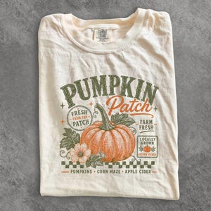 May include: A cream-colored t-shirt featuring a graphic of an orange pumpkin with green leaves and text that reads PUMPKIN Patch, FRESH FROM THE PATCH, FARM FRESH, LOCALLY GROWN, AUTUMN PICKED, PUMPKINS, CORN MAZE, and APPLE CIDER with a checkered border.