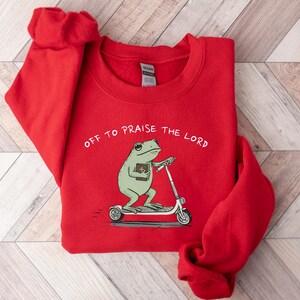 May include: A red sweatshirt with the text "OFF TO PRAISE THE LORD." The graphic features a green frog riding a scooter while holding a book. The sweatshirt is made of a soft material.