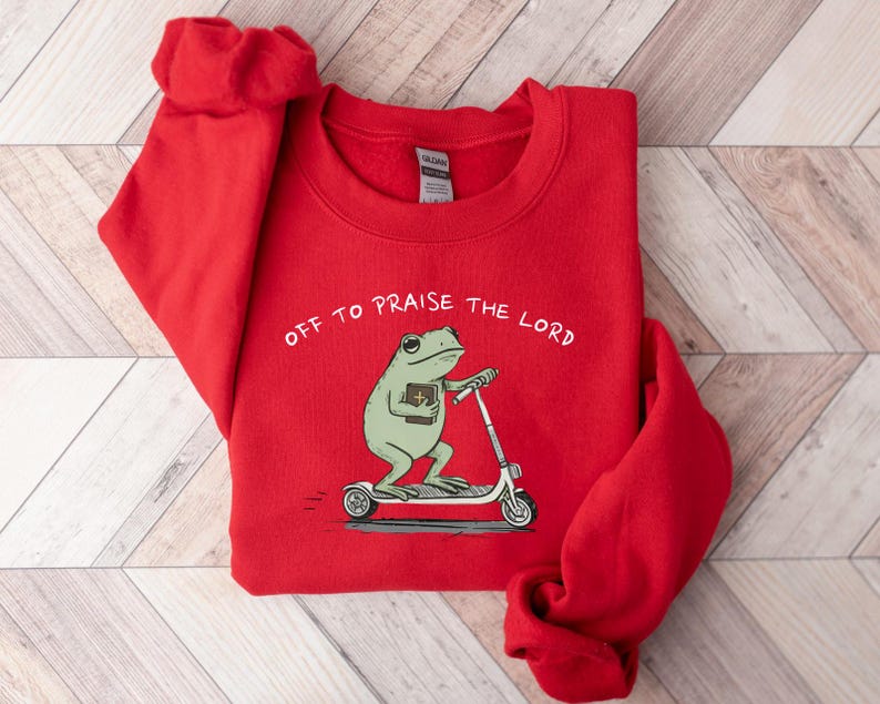 May include: A red sweatshirt with the text "OFF TO PRAISE THE LORD." The graphic features a green frog riding a scooter while holding a book. The sweatshirt is made of a soft material.