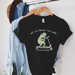 May include: Black t-shirt featuring a cartoon frog riding a scooter and holding a book. The text "OFF TO PRAISE THE LORD" is printed above the frog. The shirt is hanging on a wooden hanger.