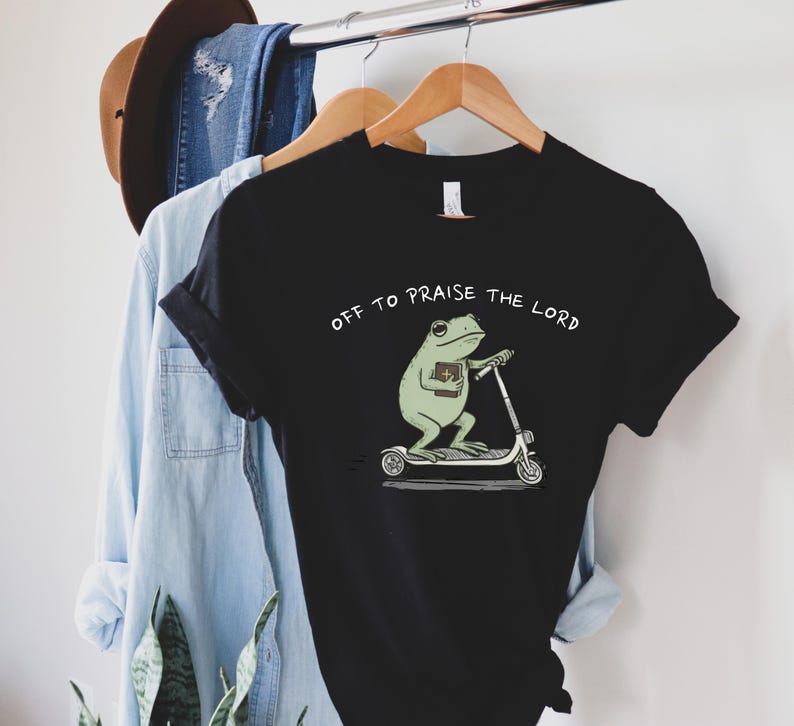 May include: Black t-shirt featuring a cartoon frog riding a scooter and holding a book. The text "OFF TO PRAISE THE LORD" is printed above the frog. The shirt is hanging on a wooden hanger.
