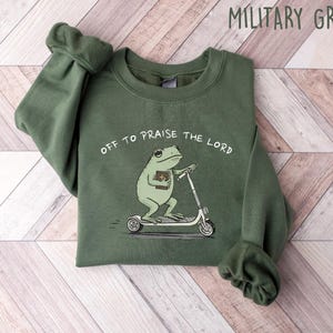 May include: Military green sweatshirt with the text "Off to Praise the Lord." The graphic features a cartoon frog riding a scooter while holding a book. The sweatshirt is made of a soft material.