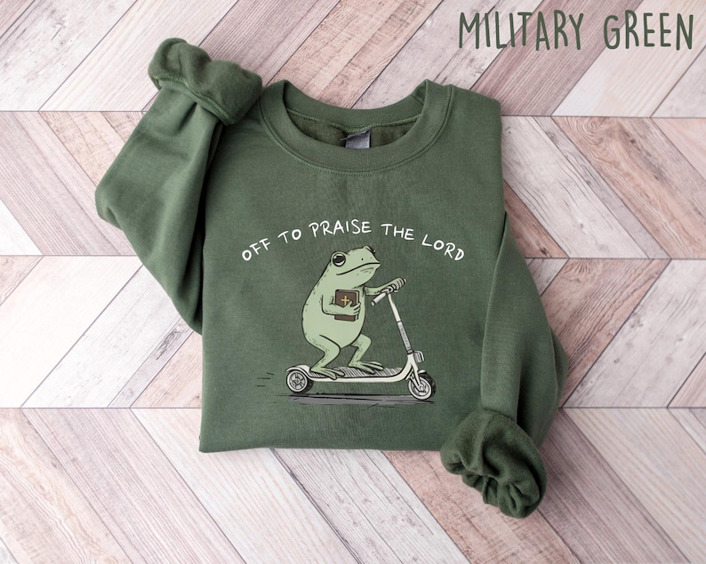 May include: Military green sweatshirt with the text "Off to Praise the Lord." The graphic features a cartoon frog riding a scooter while holding a book. The sweatshirt is made of a soft material.
