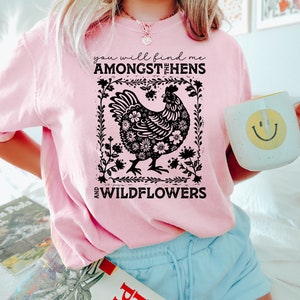 K&ouml;nnte beinhalten: Ein pinkfarbenes T-Shirt mit einer schwarzen Grafik einer Henne, die von Blumen umgeben ist. Der Text auf dem Shirt lautet "You will find me amongst the hens and wildflowers."