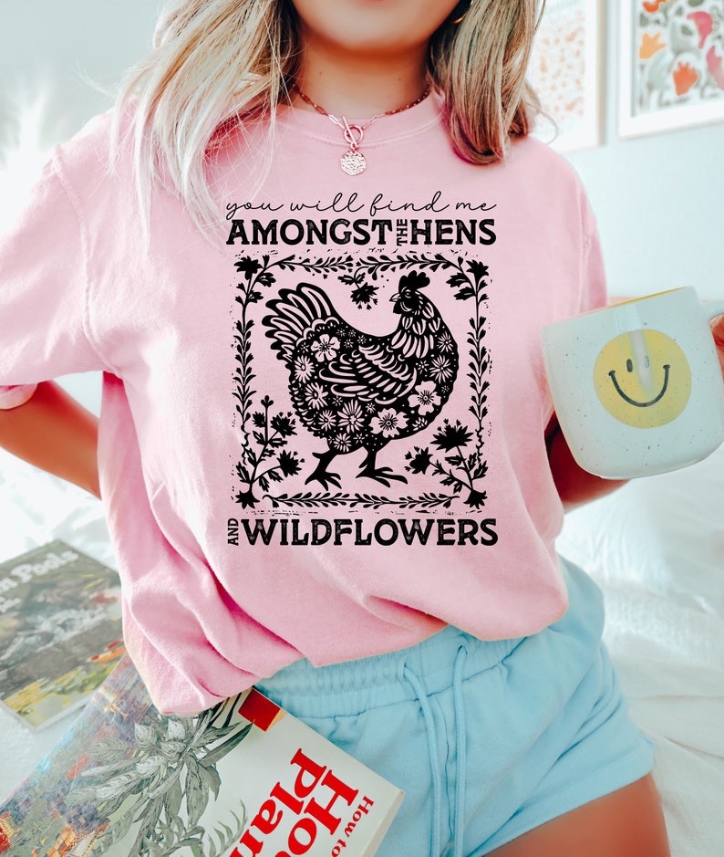 K&ouml;nnte beinhalten: Ein pinkfarbenes T-Shirt mit einer schwarzen Grafik einer Henne, die von Blumen umgeben ist. Der Text auf dem Shirt lautet "You will find me amongst the hens and wildflowers."