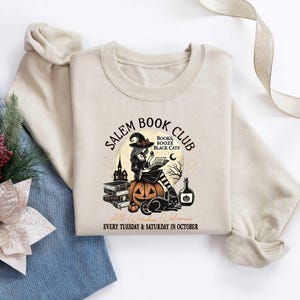 May include: A tan sweatshirt with the text "Salem Book Club" in black lettering. The text is surrounded by a witch holding a book, a pumpkin, and black cats. The text "Books, Booze, Black Cats" is written in smaller lettering below the witch. The text "Every Tuesday & Saturday in October" is written in smaller lettering below the image.