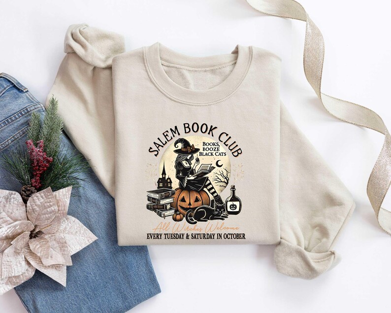 May include: A tan sweatshirt with the text "Salem Book Club" in black lettering. The text is surrounded by a witch holding a book, a pumpkin, and black cats. The text "Books, Booze, Black Cats" is written in smaller lettering below the witch. The text "Every Tuesday & Saturday in October" is written in smaller lettering below the image.