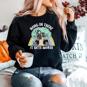 May include: Black sweatshirt featuring a graphic of a skeleton sitting on a tree with a cat. The text on the sweatshirt reads "Hang in there it gets worse".