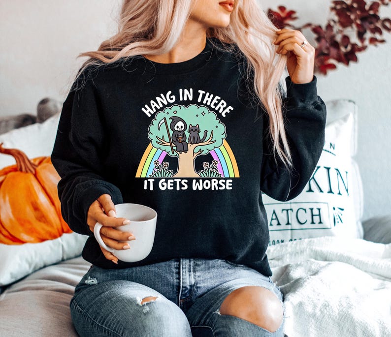 May include: Black sweatshirt featuring a graphic of a skeleton sitting on a tree with a cat. The text on the sweatshirt reads "Hang in there it gets worse".