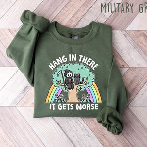 May include: A green sweatshirt with the text "Hang in there it gets worse" printed on the front. The text is surrounded by a rainbow and a cartoon image of a grim reaper and a cat sitting on a tree.