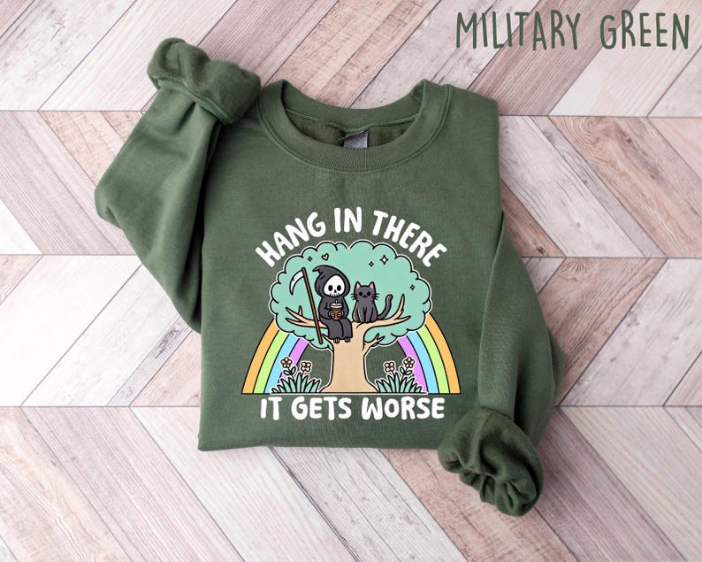 May include: A green sweatshirt with the text "Hang in there it gets worse" printed on the front. The text is surrounded by a rainbow and a cartoon image of a grim reaper and a cat sitting on a tree.