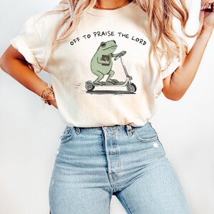 May include: Cream-colored t-shirt with the text "OFF TO PRAISE THE LORD." The shirt features a cartoon frog riding a scooter and holding a book. The frog is green, and the scooter is silver.