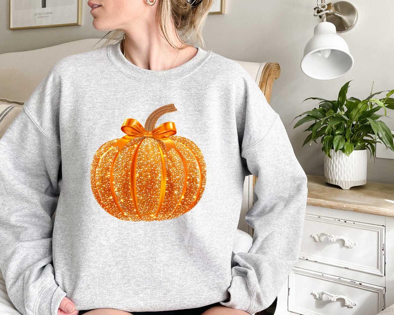 May include: A gray sweatshirt with a sparkly orange pumpkin design. The pumpkin has a brown stem and an orange ribbon.