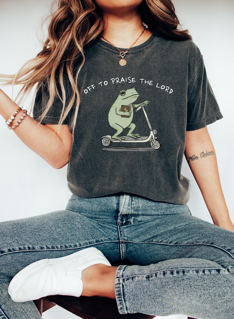 May include: Dark gray t-shirt with the text "OFF TO PRAISE THE LORD". The shirt features a green frog riding a scooter and holding a book. The frog is the central design element of this casual tee.