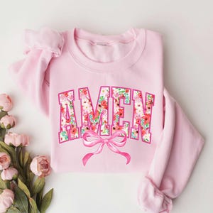 May include: Pink sweatshirt with a floral print "AMEN" design and a pink bow.