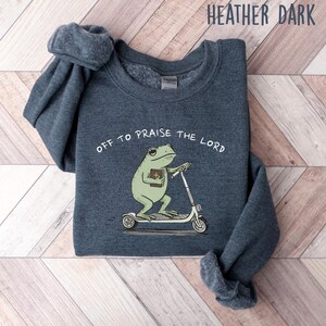 May include: Heather dark gray sweatshirt with the text "OFF TO PRAISE THE LORD." The graphic features a green frog riding a scooter and holding a book. The sweatshirt is made of a soft material.