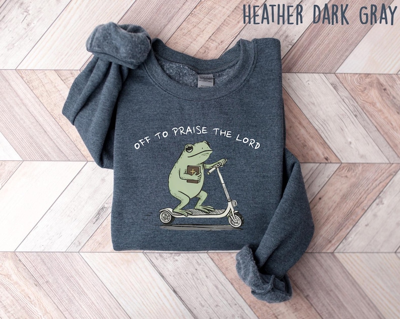 May include: Heather dark gray sweatshirt with the text "OFF TO PRAISE THE LORD." The graphic features a green frog riding a scooter and holding a book. The sweatshirt is made of a soft material.