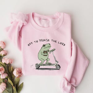 May include: A light pink sweatshirt with the phrase "OFF TO PRAISE THE LORD" printed above a cartoon frog riding a scooter and holding a book. The frog is green, and the scooter is white and gray.