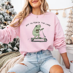 May include: A light pink sweatshirt with the phrase "OFF TO PRAISE THE LORD" printed across the top. The graphic features a green frog riding a scooter and holding a book. The sweatshirt is long-sleeved and has a crew neck.