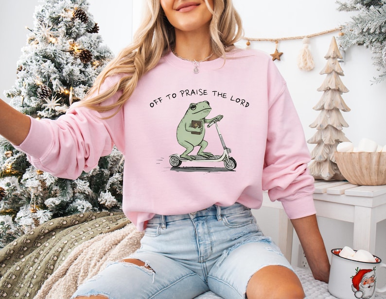 May include: A light pink sweatshirt with the phrase "OFF TO PRAISE THE LORD" printed across the top. The graphic features a green frog riding a scooter and holding a book. The sweatshirt is long-sleeved and has a crew neck.