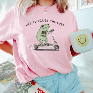 May include: A light pink sweatshirt with the phrase "OFF TO PRAISE THE LORD" printed on the front. The graphic features a green frog riding a scooter and holding a book. A white mug with a yellow smiley face is held in the hand.