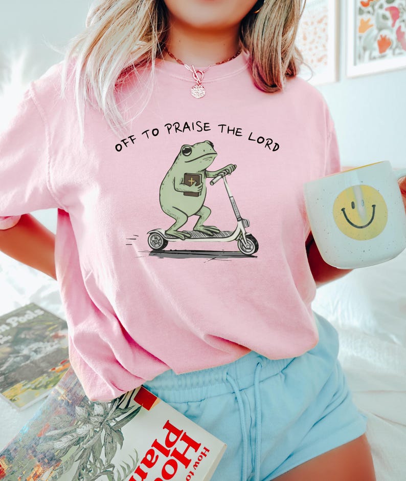 May include: A light pink sweatshirt with the phrase "OFF TO PRAISE THE LORD" printed on the front. The graphic features a green frog riding a scooter and holding a book. A white mug with a yellow smiley face is held in the hand.