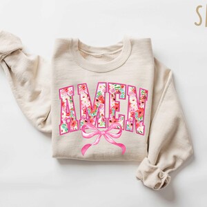 May include: A beige sweatshirt with a floral print "AMEN" graphic and a pink bow.